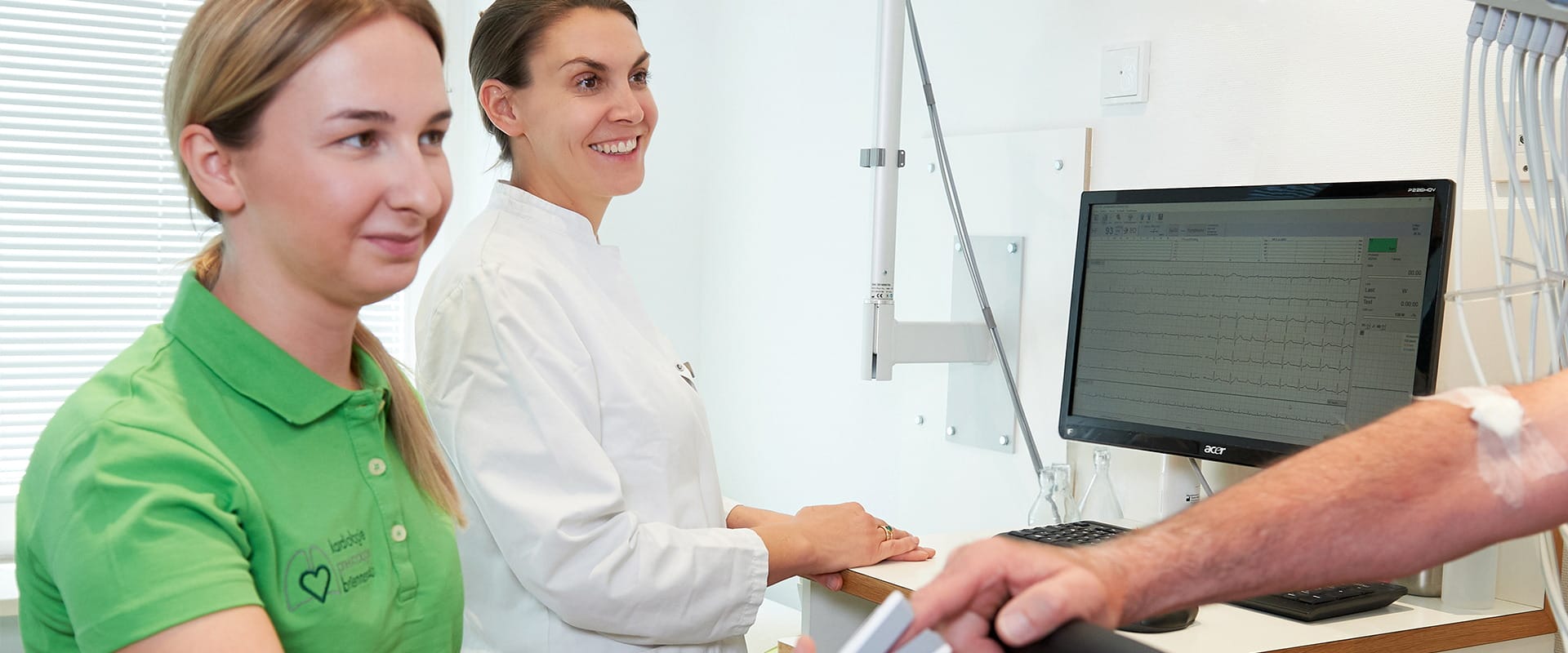 Zwei Mitarbeiterinnen der Praxis beobachten am Monitor die Kurven eines Belastungs-EKGs, rechts der Patient am Ergometer.