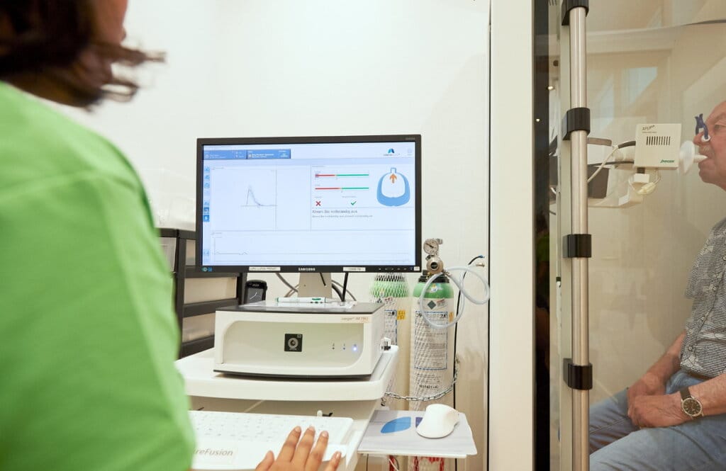 Medizinische Fachangestellte startet am Bildschirm eine Bodyplethysmographie, im Hintergrund atmet ein Patient in der Messkabine über ein Mundstück