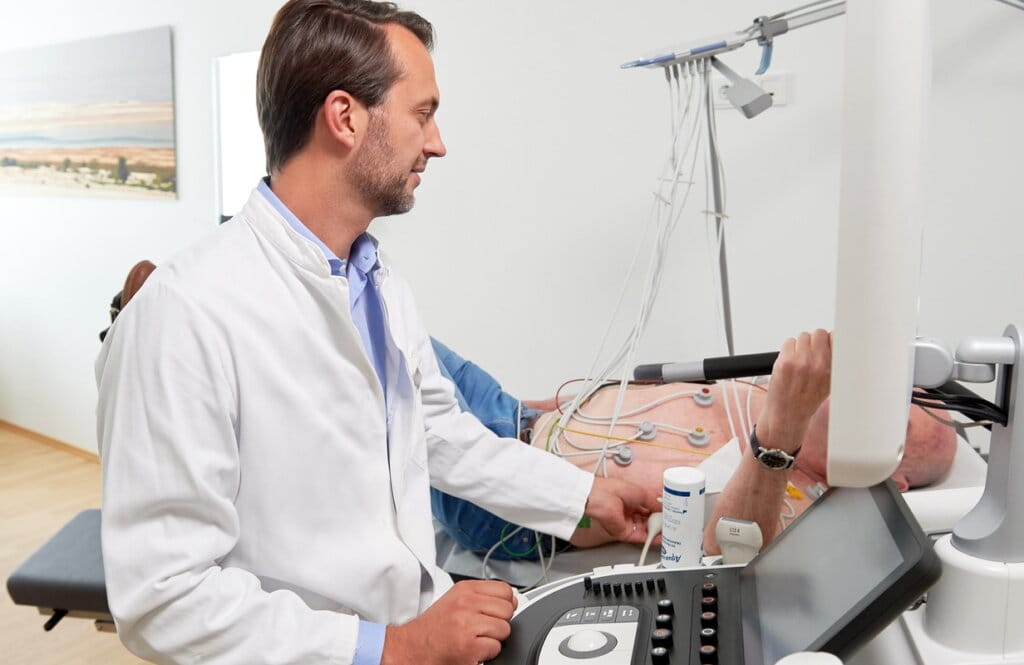 Kardiologe führt eine Echokardiographie durch, der Patient liegt mit EKG-Elektroden auf der Untersuchungsliege