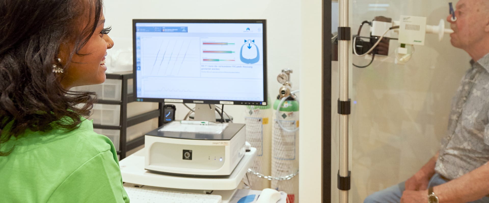 Mitarbeiterin der Praxis am Monitor mit den Messkurven einer Lungenfunktionsprüfung, rechts ein Patient in der Messkabine.